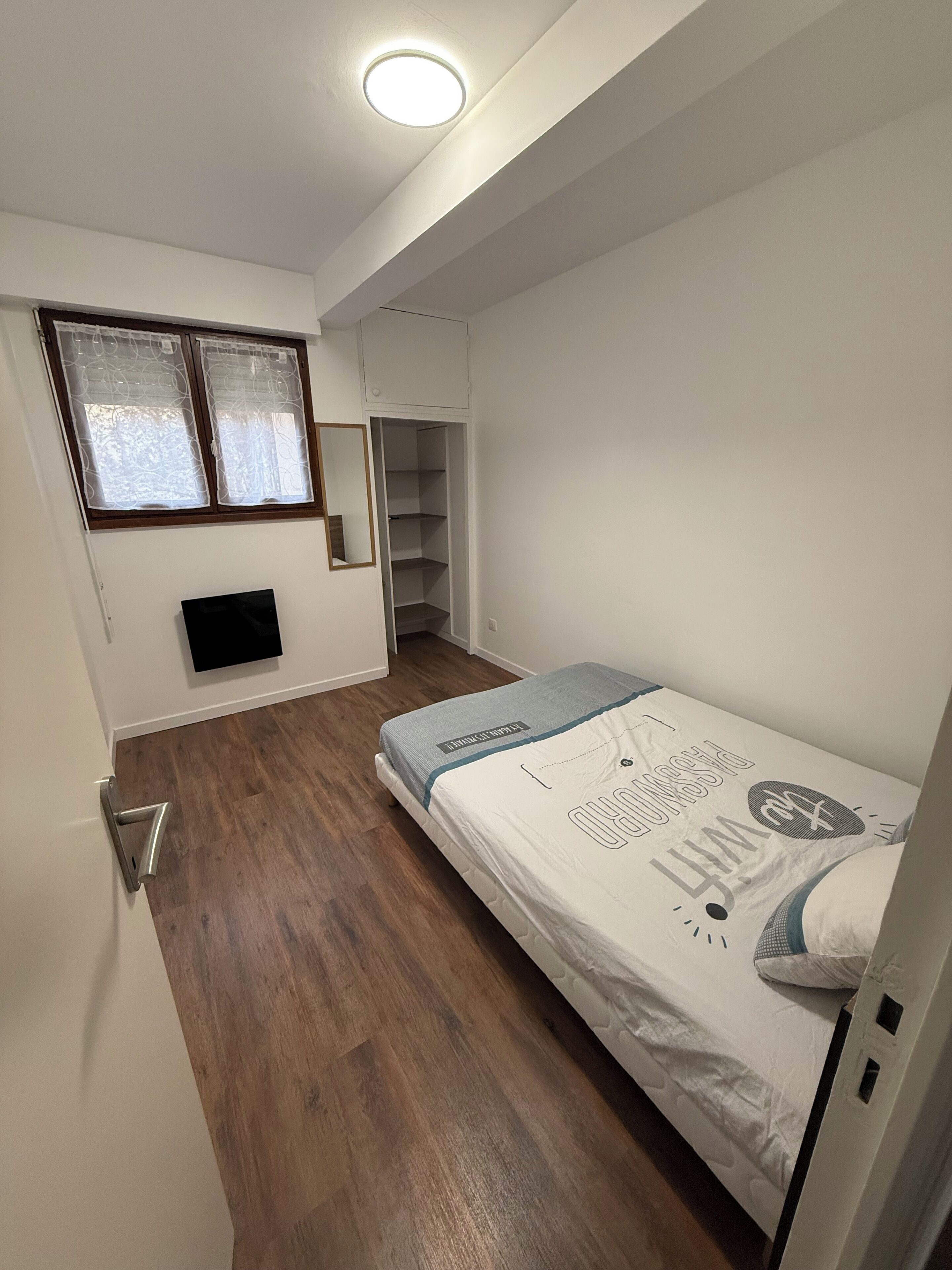 Ein Schlafzimmer mit Bett, Kleiderschrank, Spiegel und einem Fenster mit Vorhängen.