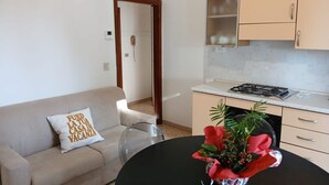 Un salon comprenant un canapé, une table basse, un coin cuisine avec des placards et une cuisinière, ainsi qu’une porte donnant sur une autre pièce.