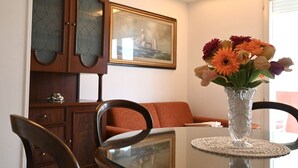 Une table à manger avec un plateau en verre transparent, un vase rempli de fleurs, un tableau encadré accroché au mur et un meuble en bois.