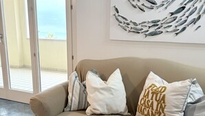 Un canapé beige agrémenté de coussins blancs et gris, une illustration représentant des poissons accrochée au mur et une porte vitrée donnant sur un balcon.