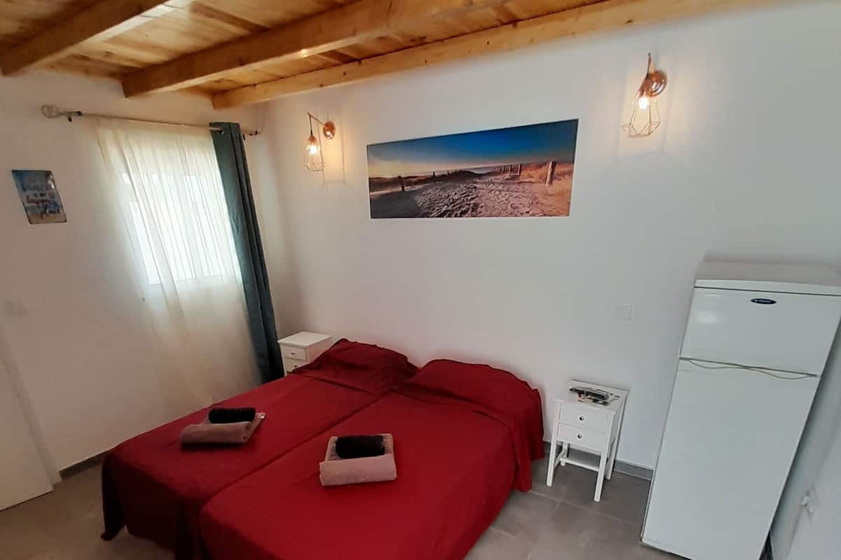 A room with two beds, a small table, a refrigerator, and a window with curtains.
