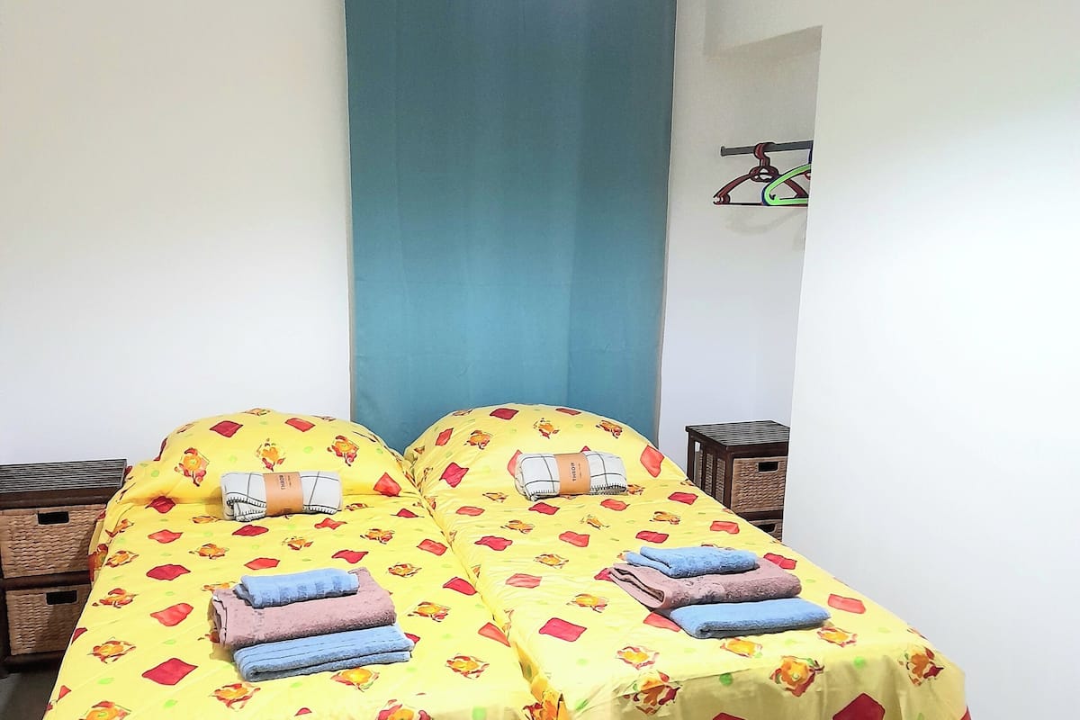 A room with two beds covered in yellow bedsheets with red and green patterns, a blue curtain, a hanging light fixture, and a small table with a basket.