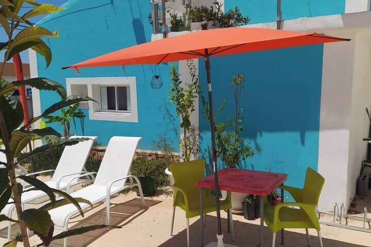 A patio area with a blue building, white lounge chairs, a red umbrella, and a small table with chairs.