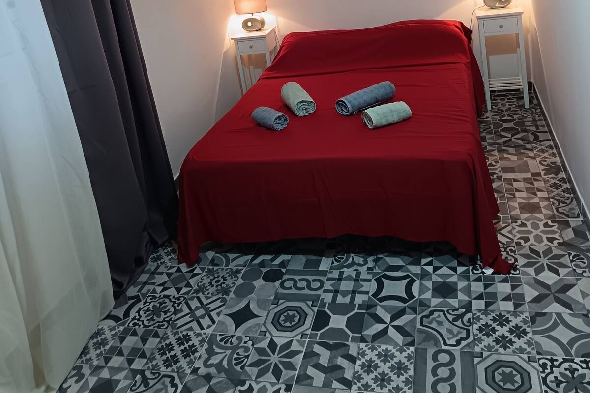 A hotel room with a red bedspread, two bedside lamps, a large floral painting on the wall, and patterned floor tiles.