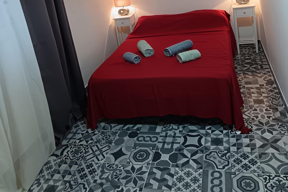 A hotel room with a red bedspread, two bedside lamps, a large floral painting on the wall, and patterned floor tiles.