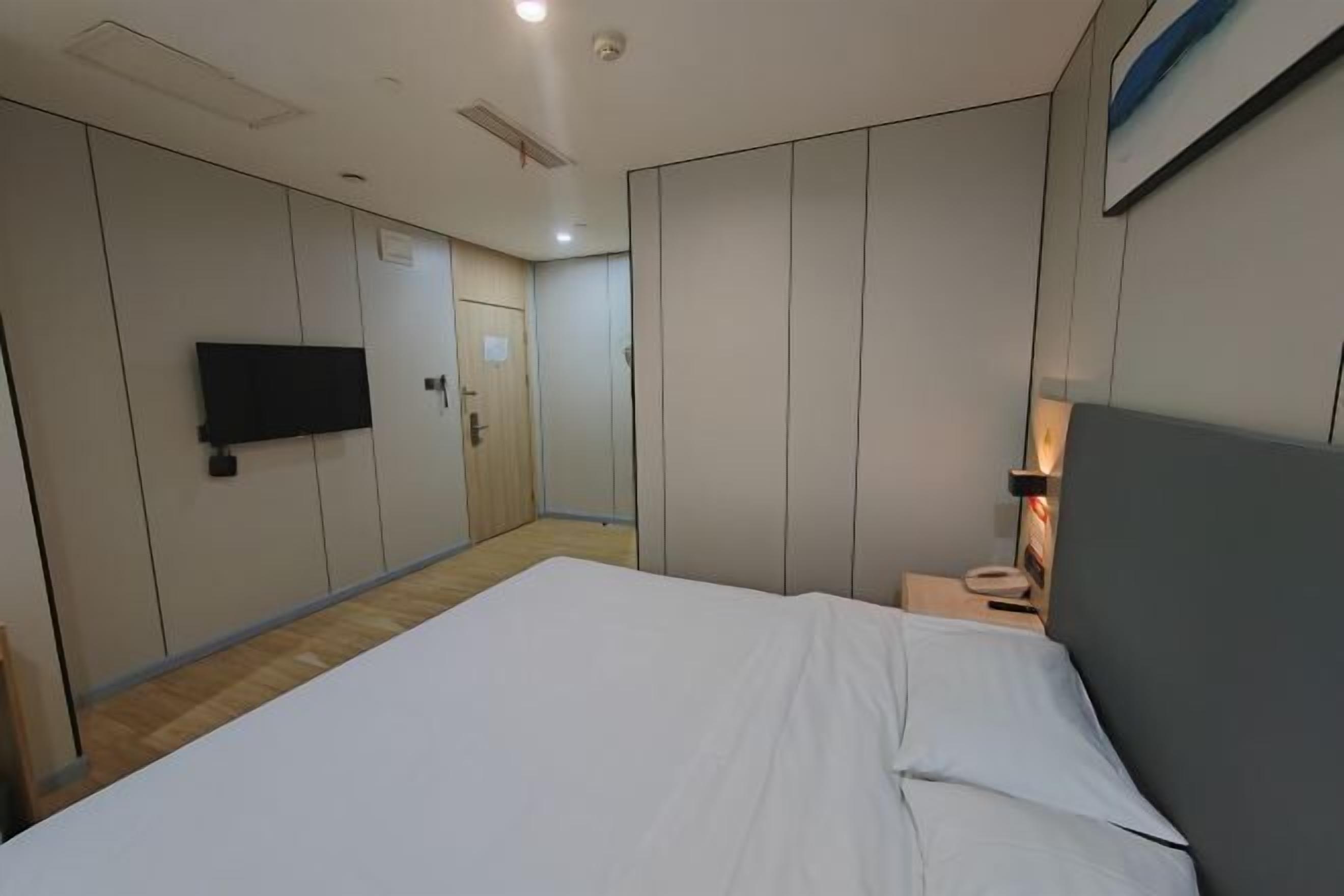 A hotel room with a large bed, a flat-screen TV mounted on the wall, a bedside table with a lamp, and a door leading to another room.
