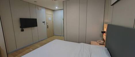 A hotel room with a large bed, a flat-screen TV mounted on the wall, a bedside table with a lamp, and a door leading to another room.