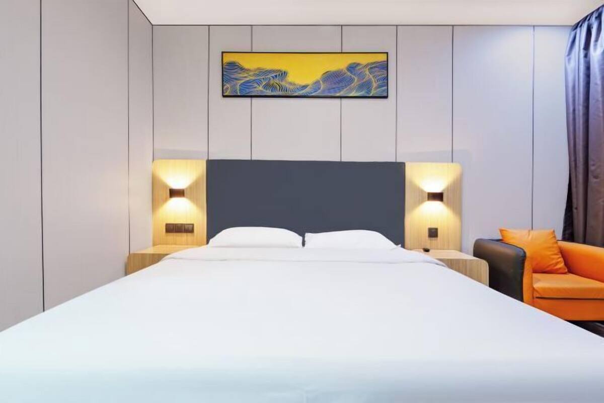 A modern hotel room with a large bed, two bedside lamps, a painting on the wall, and an orange chair.