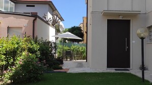 Ein Wohngebäude mit einer schwarzen Tür, einem kleinen Garten mit Rasen und einer Terrasse mit Tisch und Stühlen.