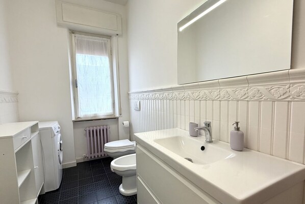 A bathroom with a white sink, toilet, and washing machine, a window with a curtain, and a radiator.