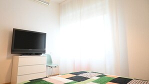 Ein Zimmer mit einem Flachbildfernseher auf einer weißen Kommode, einer Bettdecke auf dem Bett und einem Fenster mit durchsichtigen Vorhängen.