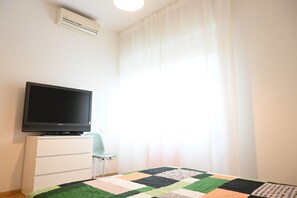 A room with a flat-screen TV on a white dresser, a quilt on the bed, and a window with sheer curtains.