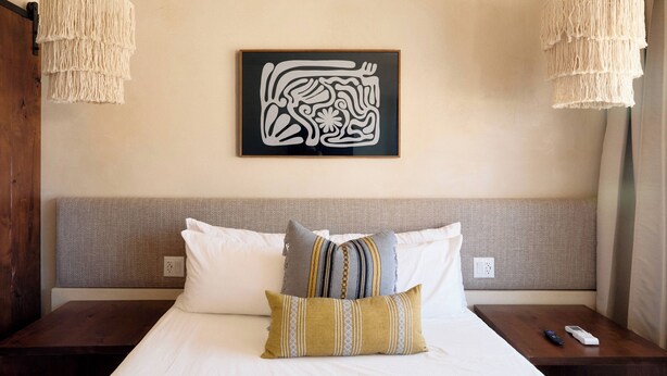 Une chambre d’hôtel comprenant un lit, deux tables de chevet, une œuvre d’art accrochée au mur et deux luminaires suspendus.