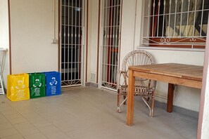 Una habitación con una mesa de madera y una silla de mimbre, una ventana con barrotes y bolsas de reciclaje en el suelo.