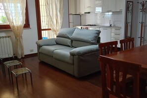 Un salon avec un canapé, deux fauteuils, une table à manger et, au fond, une cuisine.