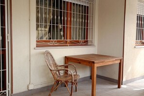 Una habitación con una mesa de madera y una silla de mimbre, una ventana con barrotes y una pared sencilla.