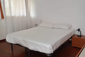 Un lit simple avec un matelas et des oreillers blancs, une table de chevet avec une lampe et une fenêtre avec des rideaux.