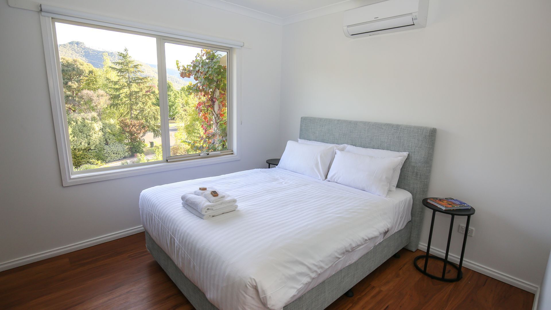A neatly made bed with white linens and a folded towel, a window with a view of trees, and a small table with a book.