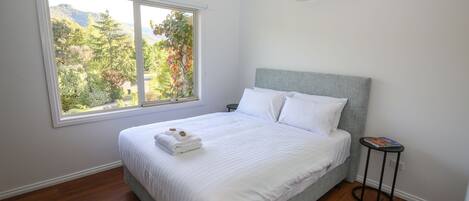 A neatly made bed with white linens and a folded towel, a window with a view of trees, and a small table with a book.