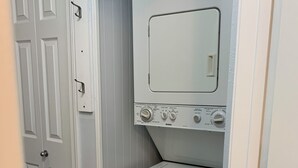 A compact laundry room with a front-loading washer and dryer, a door with a handle, and a tiled floor. - Studio in The Heart of Soldotna (Soldotna)