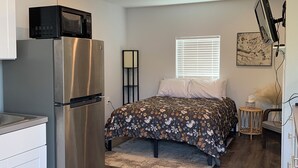 A compact bedroom with a bed, a built-in refrigerator, a microwave, a bedside table, and a window with blinds. - Studio in The Heart of Soldotna (Soldotna)