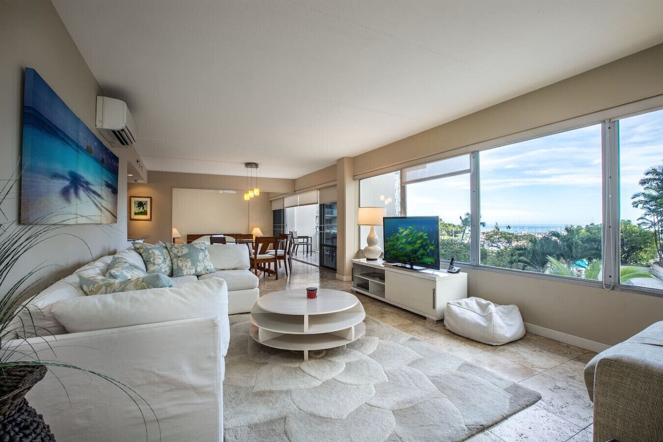 A modern living room with a white sofa, a coffee table, and a TV. There is a large window with a view of the ocean.