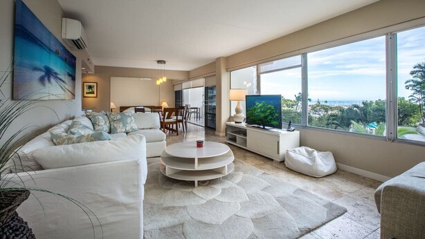Ein modernes Wohnzimmer mit einem weißen Sofa, einem Couchtisch und einem Fernseher. Ein großes Fenster bietet Blick aufs Meer.