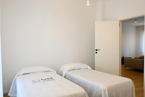 Une chambre avec deux lits simples, une porte donnant sur une autre pièce et un interrupteur visible sur le mur.