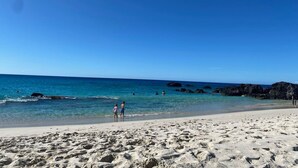 A beach with clear water, rocky outcrops, and people walking along the shore.