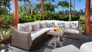 A covered outdoor seating area with a wicker sofa and a wooden table, surrounded by palm trees and tropical plants.