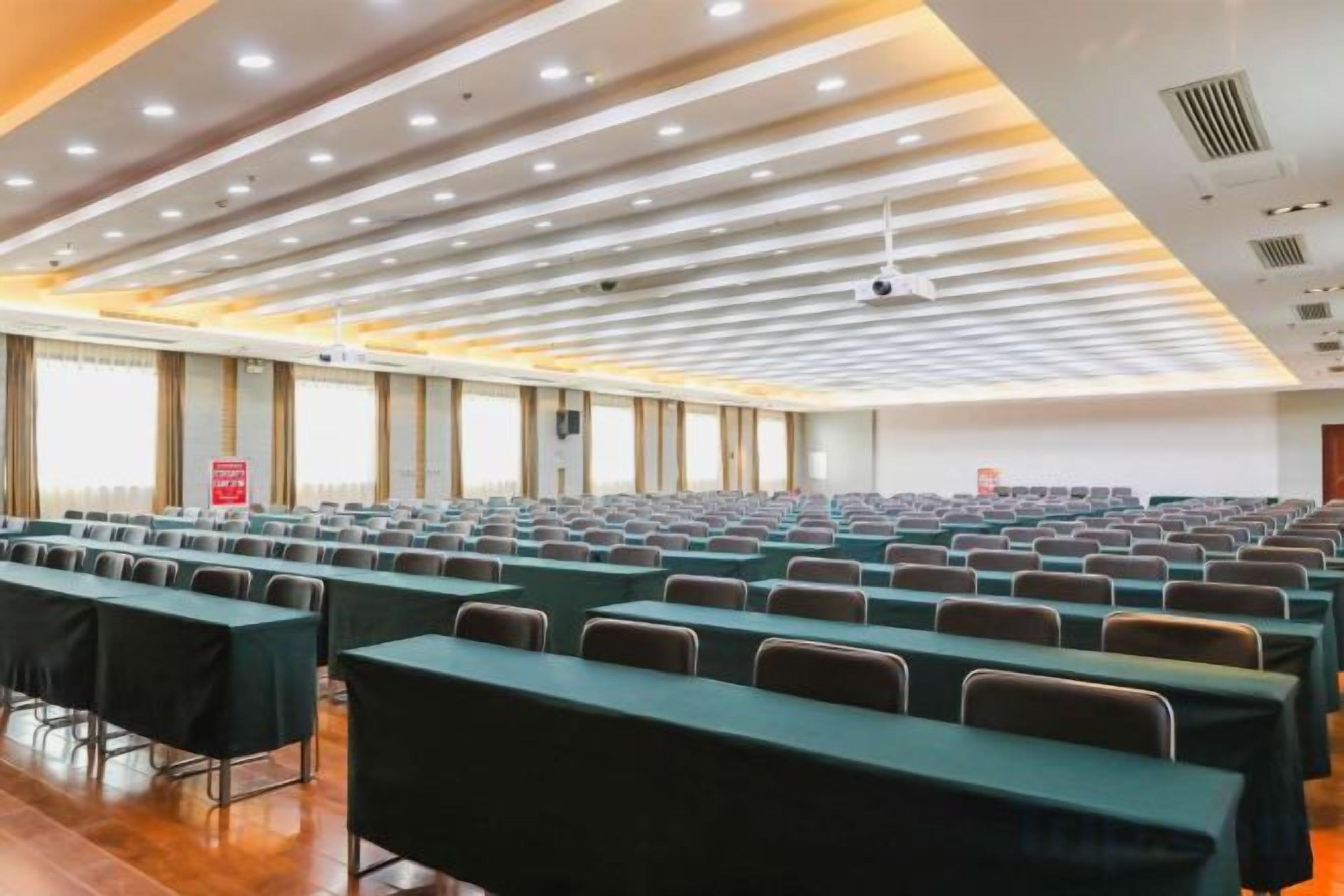 A conference room with rows of tables and chairs, a ceiling with recessed lighting, and a projector screen at the front.
