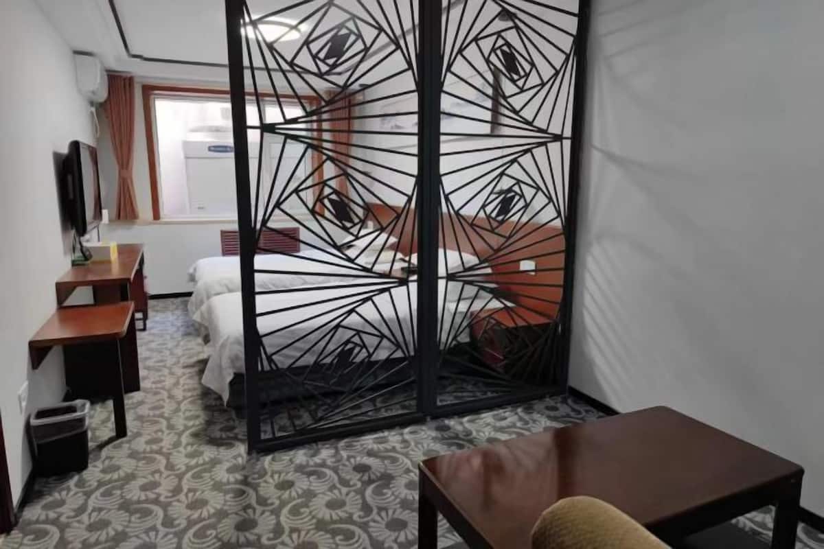 Une chambre d’hôtel comprenant un lit, un bureau, une chaise et un élément de séparation décoratif orné de motifs géométriques.