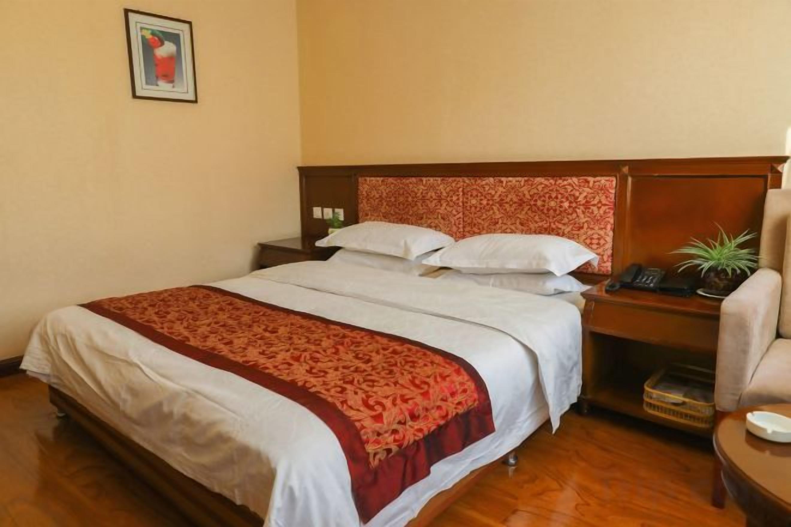 Habitación de hotel con una cama grande, un cabecero de madera, una mesita de noche con una lámpara, una silla y una pequeña mesa con una planta.