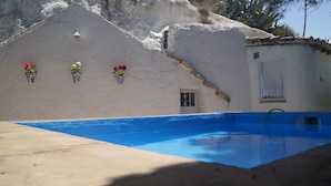 A swimming pool with a blue interior, surrounded by a concrete deck. There is a white building with a window and a door, and three flower pots with red flowers on the wall. - Cave House "La Estrella" 10 minutes from Granada center. (Monachil)