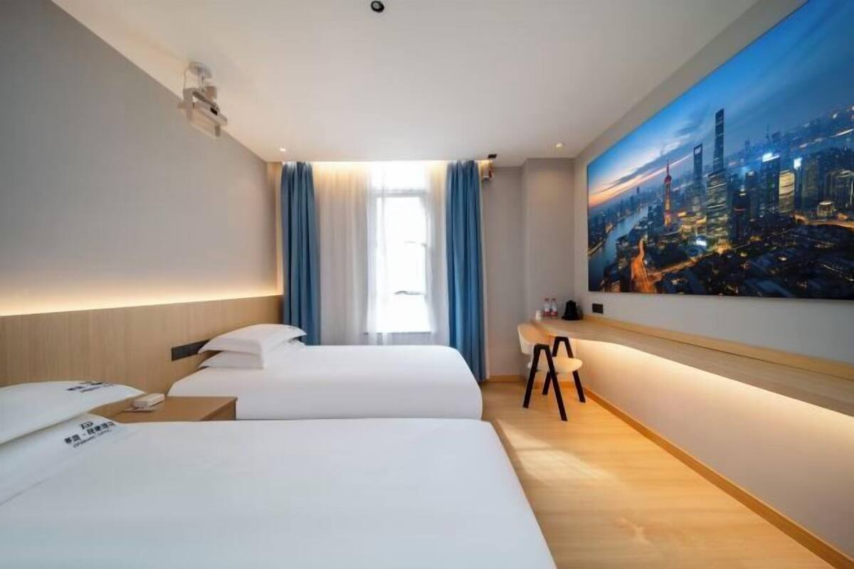 A hotel room with two beds, a desk, a large cityscape painting, and a window with curtains.