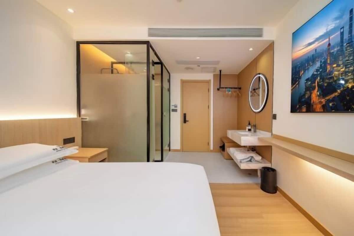 A hotel room with a bed, a glass-enclosed shower, a sink, and a cityscape painting.