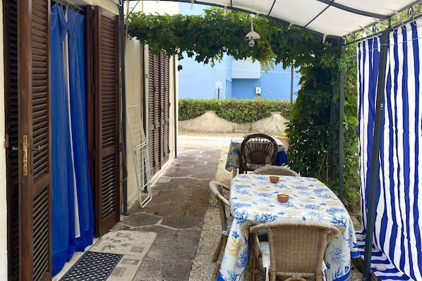 An outdoor patio with a table and chairs, a covered area with a blue and white striped curtain, and a view of a blue building in the background.