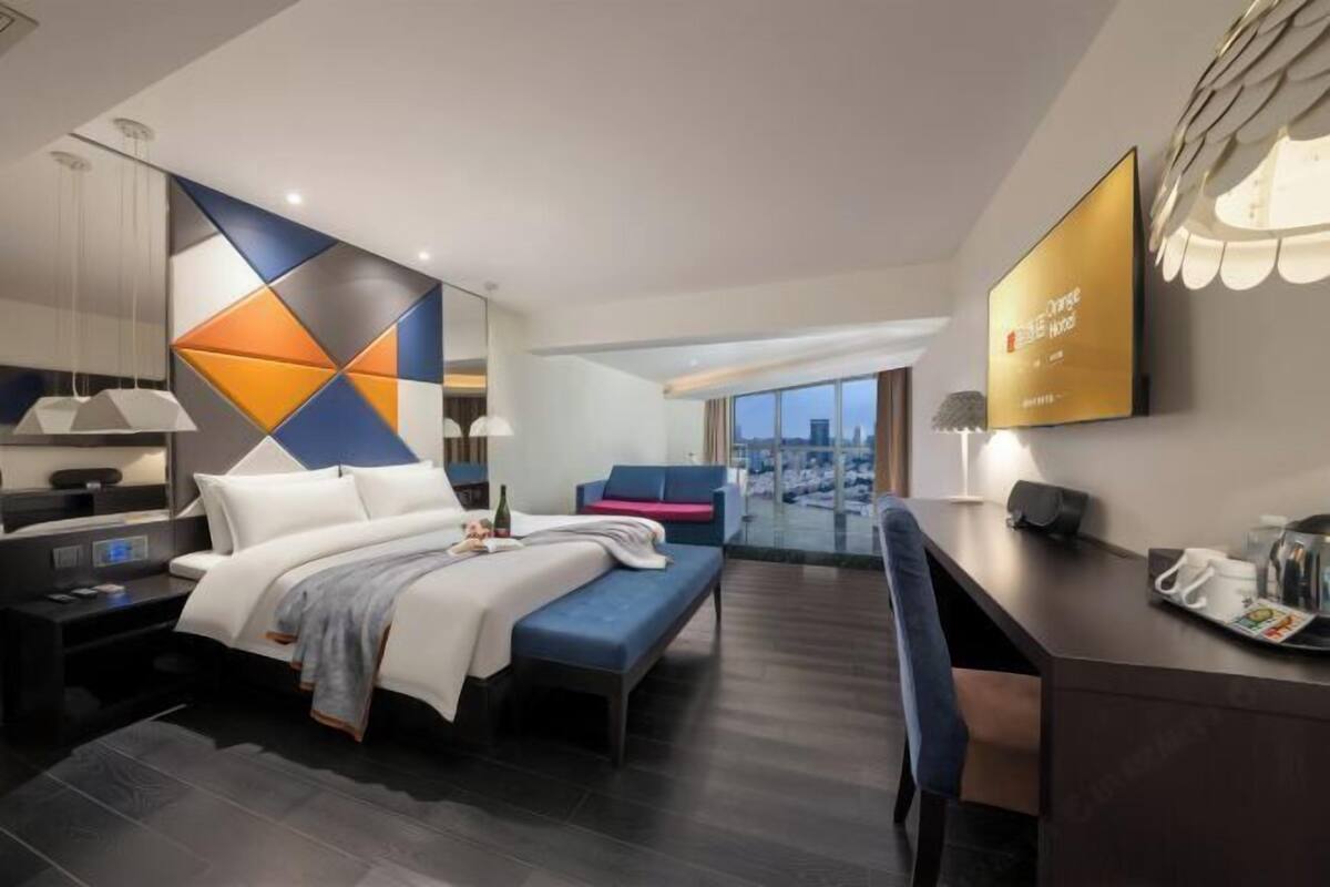 A modern hotel room with a bed, bedside table, desk, and a large window offering a city view.