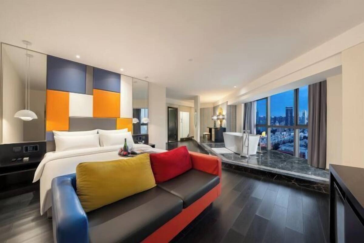 A modern hotel room with a bed, a sofa with colorful cushions, a desk, and a large window offering a city view.