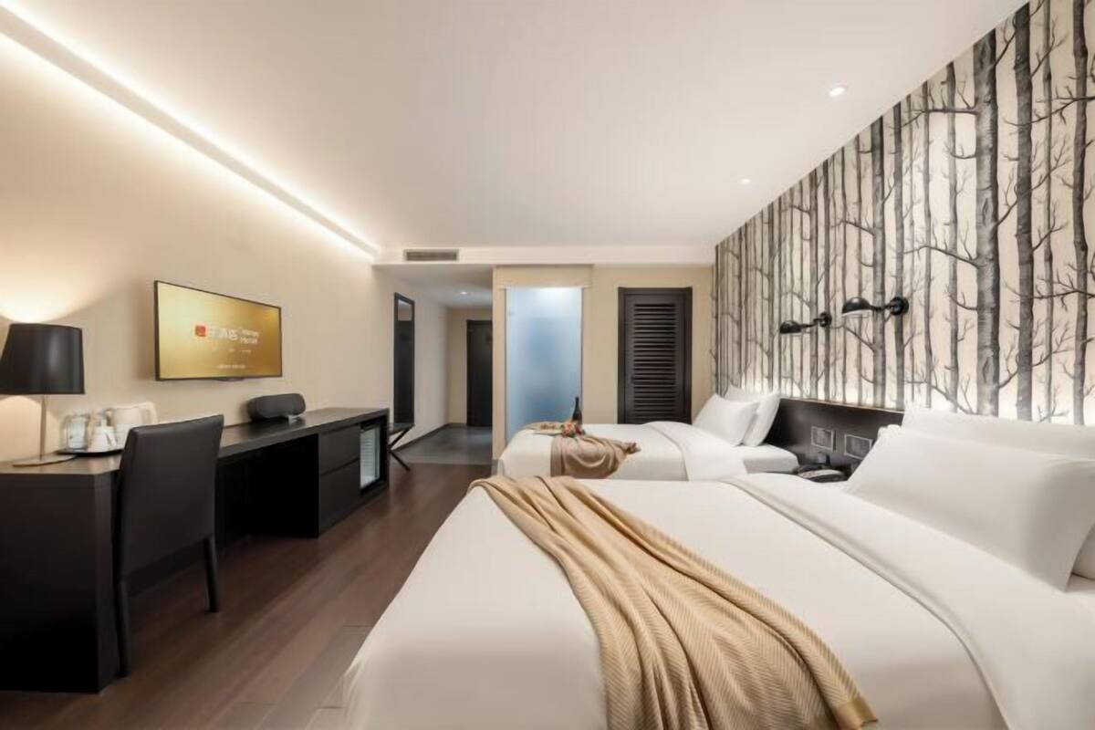 A hotel room with a bed, a desk, a chair, a TV, and a wall with a tree pattern.