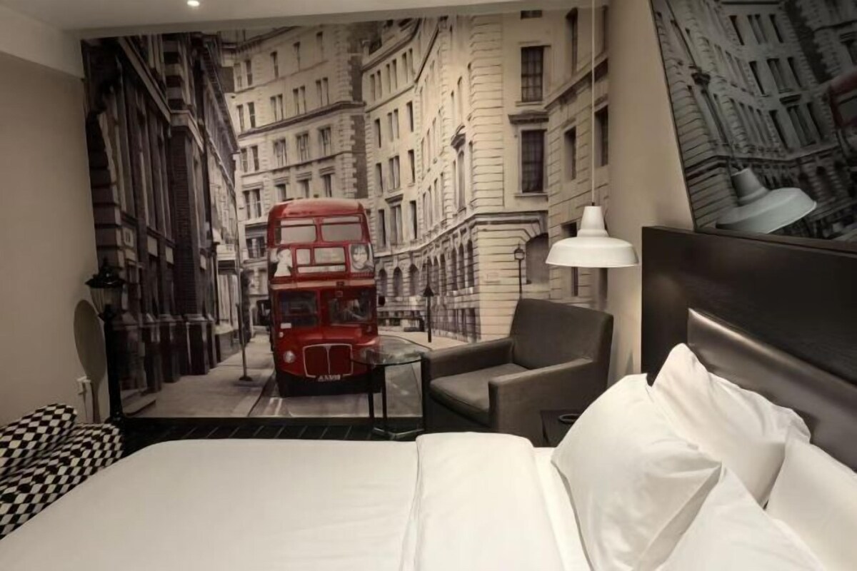A hotel room with a large bed, a chair, a table, and a red double-decker bus mural on the wall.