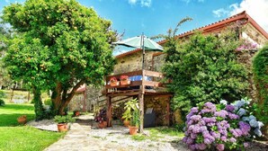 A stone pathway leading to a rustic building with a balcony, surrounded by lush greenery and flowering plants.