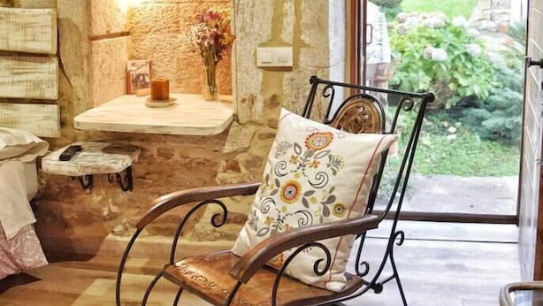A rustic interior with a stone wall, a wooden rocking chair with a patterned cushion, a small shelf with a vase, and a view of a garden through a glass door.