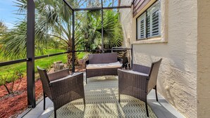 A covered patio with wicker furniture, a small table, and a view of a garden with palm trees.