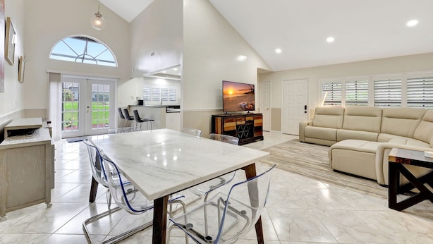 A spacious living area with a large dining table, a sofa, and a television. There is a kitchen area with cabinets and a window.