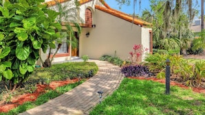 A house with a paved pathway leading to the entrance, surrounded by lush greenery and a well-maintained garden.