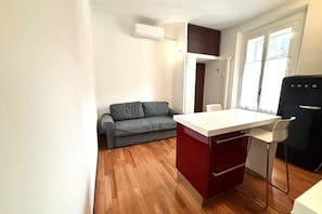 A compact kitchen with a white countertop, a red cabinet, a grey sofa, and a window with curtains.