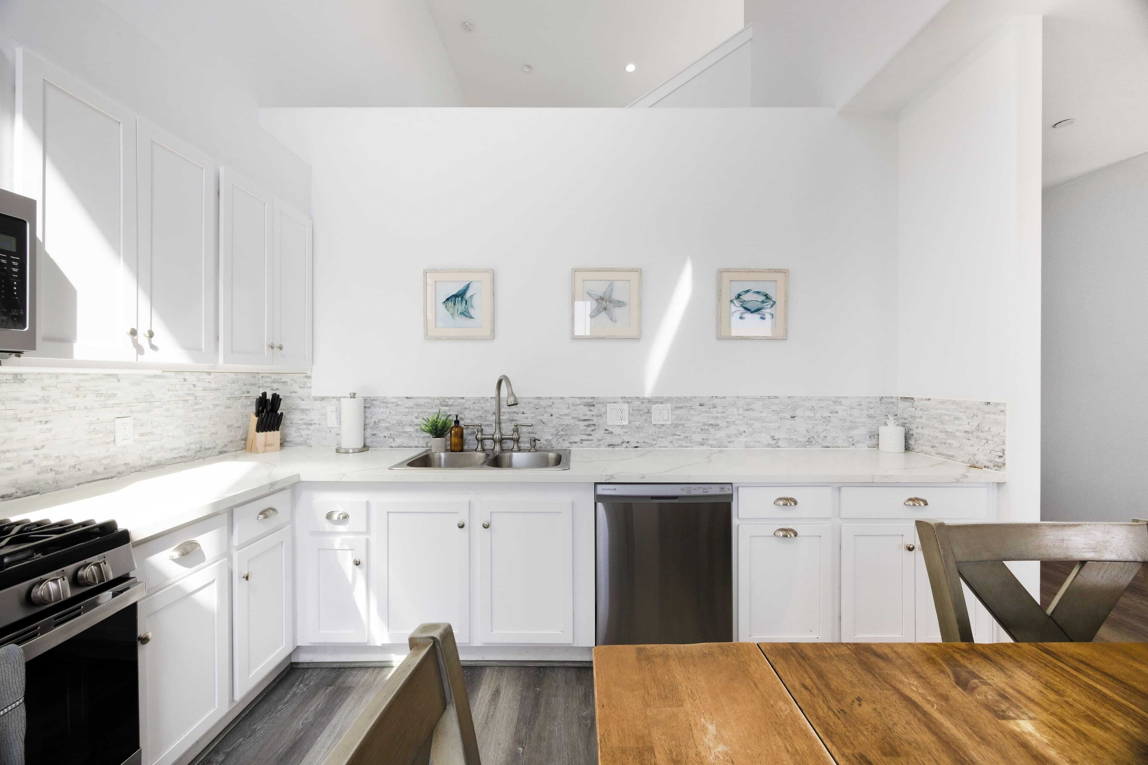 A modern kitchen with white cabinets, a marble backsplash, and stainless steel appliances. There are framed pictures on the wall and a wooden dining table.