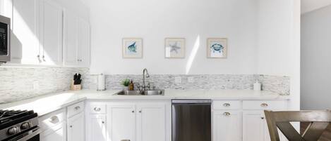 A modern kitchen with white cabinets, a marble backsplash, and stainless steel appliances. There are framed pictures on the wall and a wooden dining table.