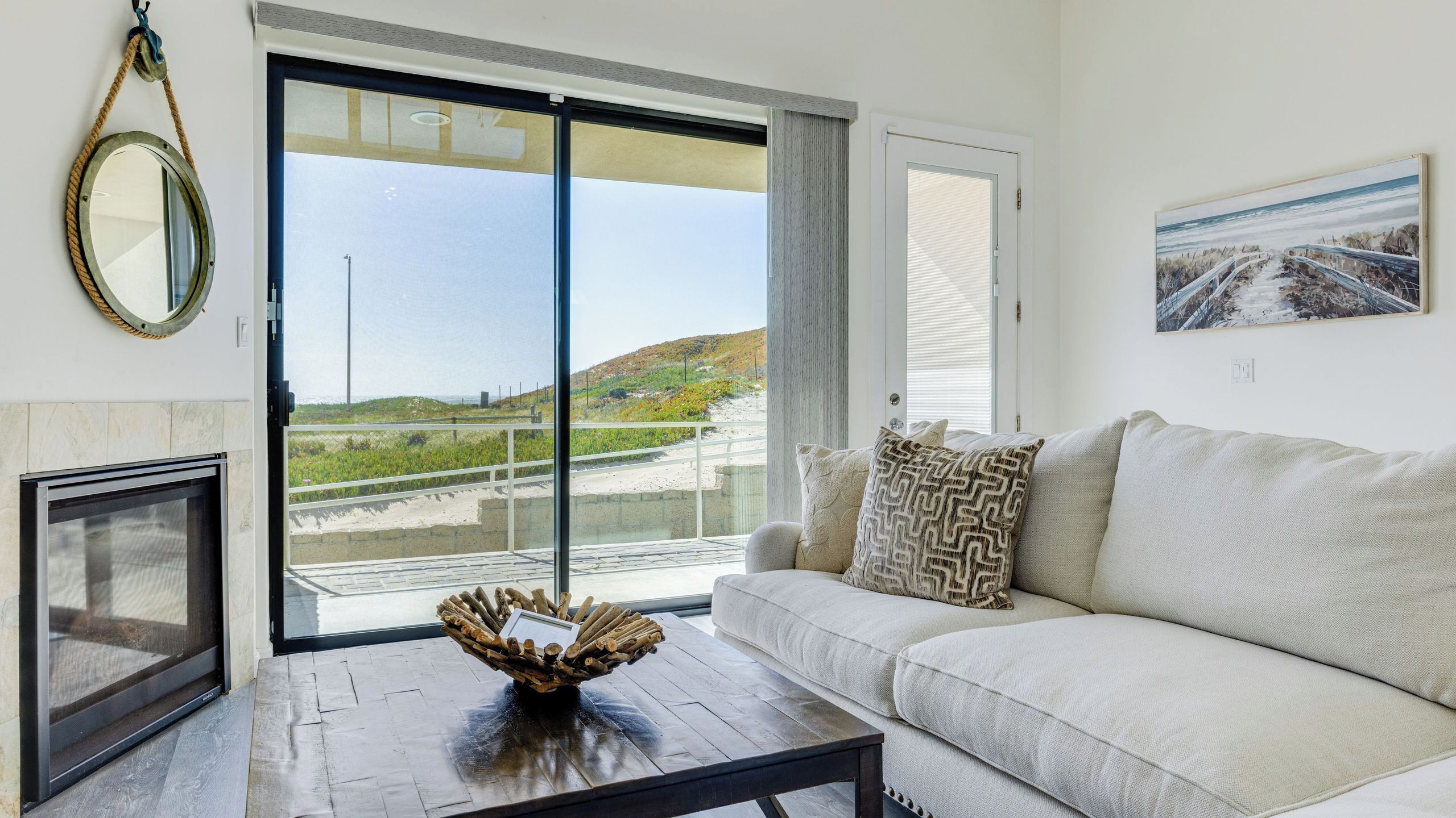 A living room with a sofa, a coffee table with a decorative bowl, a fireplace, and a large sliding glass door leading to a balcony with a view of the beach and hills.